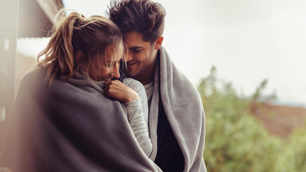 Romantic couple on a winter holiday. Man and woman standing together in a hotel room balcony wrapped in blanket. Couple embracing and smiling.