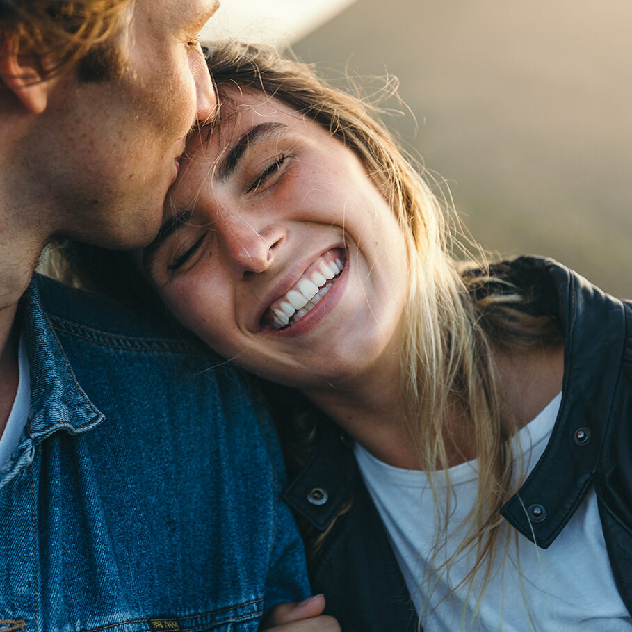 Close-up of romantic young boyfriend kissing smiling beautiful girlfriend on forehead while enjoying weekend at sunset