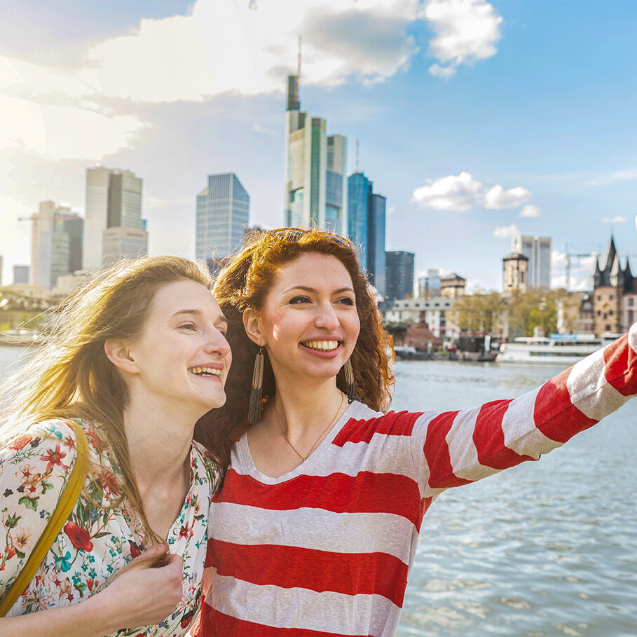 Two beautiful young women taking a selfie with Frankfurt skyline on background on a spring day. They are smiling and looking at the smart phone. Friendship and travel concepts.