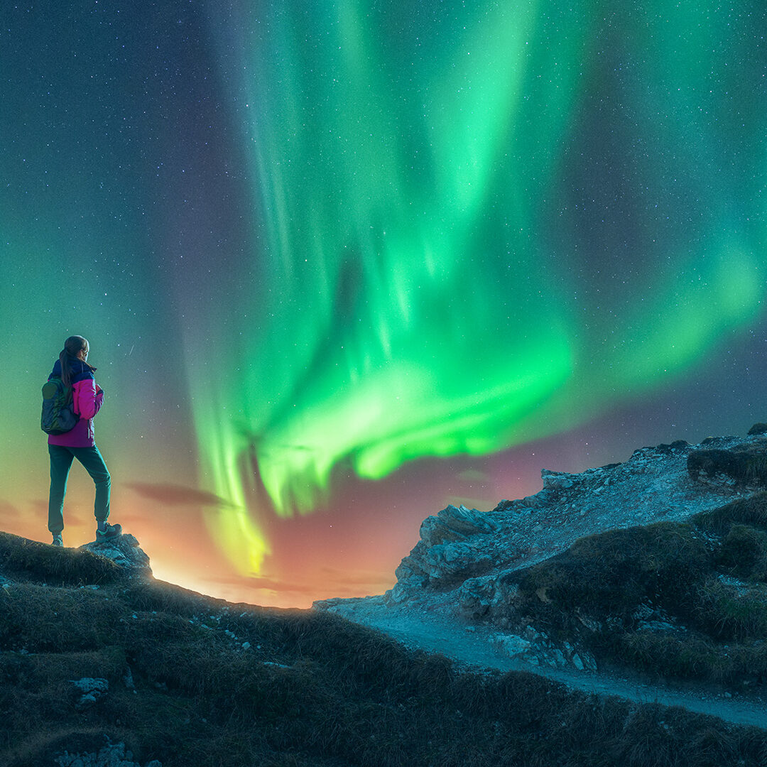 Northern lights and young woman on mountain peak at night. Aurora borealis and silhouette of alone girl on mountain trail. Landscape with polar lights. Starry sky with bright aurora. Travel background